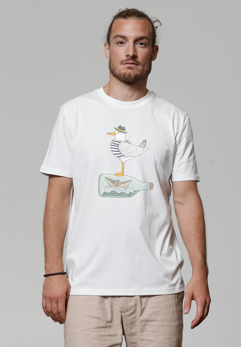 Witte katoenen t-shirt met een afbeelding van een eend in een gestreept shirt en hoed, staand op een groene glazen fles met een papieren bootje erin.