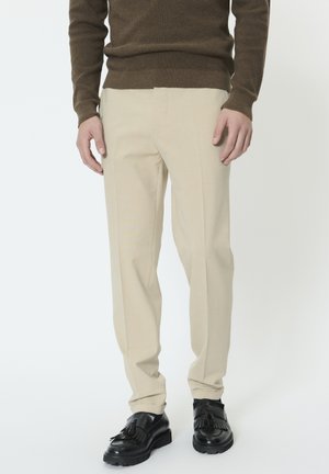 Egy férfi bézs nadrágot, fekete bojtos mokaszint és barna hosszú ujjú pulóvert visel, egyszerű háttér előtt áll.