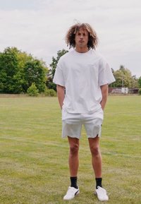 Hvit bomull t-skjorte og lyse shorts, står på gress. Hvite joggesko og svarte sokker. Krøllete hår, hender i lommer, avslappet antrekk.