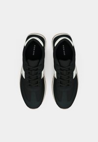 Ασπρόμαυρα αθλητικά παπούτσια με κορδόνια και καφέ σόλες, φωτoγραφημένα από πάνω σε ανοιχτό γκρι φόντο.