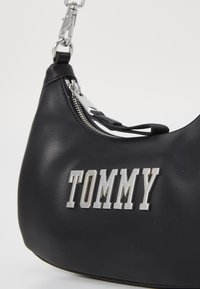 Svart lær skulderveske med en sølvfarget glidelås, gummizipperpull og en fremtredende sølvfarget "TOMMY"-logo på siden.
