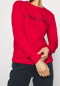 Czerwona bluza z okrągłym dekoltem i ściągaczami na mankietach, z widocznym czarnym haftem z napisem "Calvin Klein" na piersi.