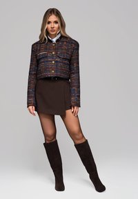 Skórzana kurtka w kratę z odcieniami niebieskiego, pomarańczowego i brązowego; dwie przednie kieszenie i złote guziki; zestawiona z brązową spódnicą i brązowymi kozakami do kolan.