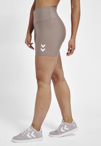 Athletic shorts in taupe material with a high waistband, featuring a white chevron logo on the side. Paired with gray sneakers.