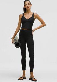 Sort crop top med V-hals og brede stropper, parret med højtaljede sorte leggings med et subtilt mønster. Ruller en yoga måtte.