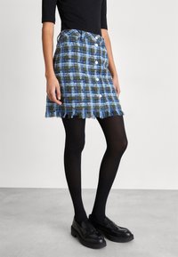 Mini falda a cuadros azul y verde con dobladillo deshilachado, cinco botones blancos en el frente, combinada con medias negras y mocasines.