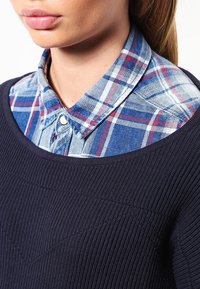 Granatowy sweter dzianinowy nałożony na niebieską i białą koszulę w kratę z guzikami. Koszula ma kołnierzyk i kontrastowe kolory w wzorze w kratkę.