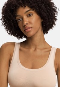 Vaaleanpunainen toppi pyöreällä kaula-aukolla ja paksuilla olkaimilla, valmistettu sileästä kankaasta. Mallilla on kiharat tummat hiukset ja hänen ihonsävy on luonnollinen.