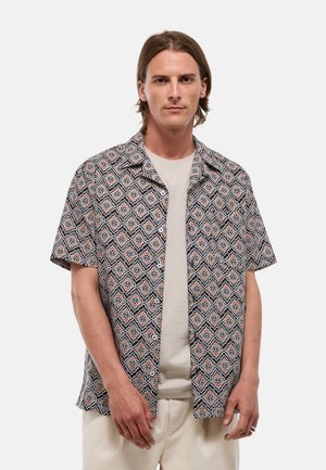 Homme aux cheveux bruns et raides, portant une chemise à manches courtes à motifs par-dessus un T-shirt beige et un pantalon clair, debout devant un fond uni.