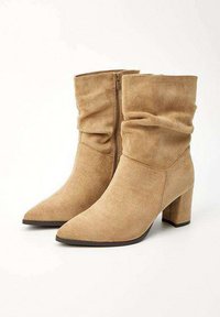 Bottines en suède beige avec un design froncé, bout pointu, talons bloc et une fermeture éclair sur le côté. Texture lisse et forme structurée.