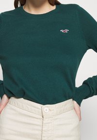 Kvinna som bär en åtsittande grön stickad tröja med en liten röd och vit fågel-logotyp, instoppad i höga beiga byxor med knappstängning.