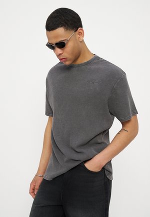 Νεαρός άνδρας με σκούρα γυαλιά ηλίου, γκρι μπλούζα με υφή, μαύρο παντελόνι, με το ένα χέρι στην τσέπη, κοιτάζοντας προς τα κάτω σε απλό λευκό φόντο.