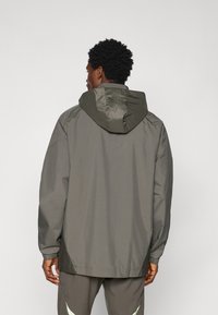 Grå, letvægtsjakke med hætte, der har elastiske manchetter og en afslappet pasform. Glat tekstur og minimalistisk design, set fra ryggen.