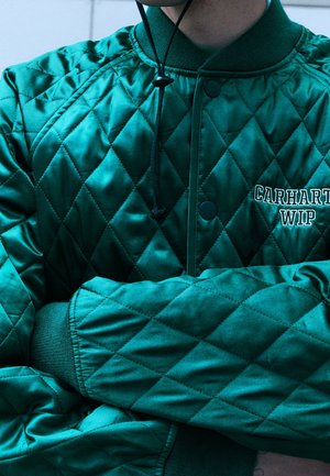 Persona che indossa una giacca trapuntata verde turchese con il logo "CARHARTT WIP" sul petto, braccia incrociate e giacca abbottonata.