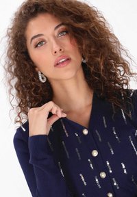 Tengerészkék kardigán flitterekkel és gyöngygombokkal. V-nyakú, bordázott mandzsettákkal rendelkezik. Anyaga puha és sima tapintású.