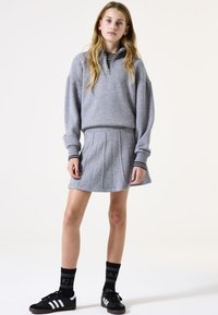 Grijze pullover met een halve rits en zwarte streepaccenten, gecombineerd met een lichtgrijze geplooide rok en zwarte sneakers met witte strepen.