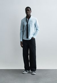 Άνδρας που φοράει ανοιχτό γαλάζιο πουκάμισο με κουμπιά και χαλαρή μαύρη γραβάτα, μαύρο παντελόνι φαρδύ από το πόδι και μαύρα και άσπρα αθλητικά παπούτσια, στέκεται μπροστά σε απλό φόντο.