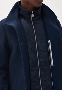 Abrigo de mezcla de lana en color navy con un alto cuello, una chaqueta interior acolchada con cremallera y un bolsillo frontal texturizado con un acento a rayas.
