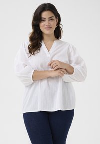 Weißes Pulloverhemd mit V-Ausschnitt, 3/4 Puffärmeln, dezenten vertikalen Streifen und gerafften Bündchen; kombiniert mit dunkelblauen Jeans.
