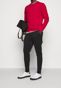 Sudadera roja, pantalones cargo negros con bolsillos laterales y zapatillas blancas con suelas negras. El material parece ser una mezcla de algodón y poliéster.