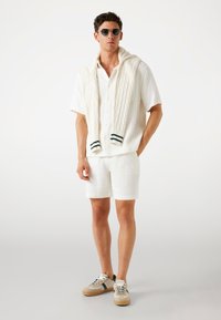 White textured short-sleeve shirt with a cable-knit sweater draped over shoulders, paired with matching shorts and sneakers in neutral tones.