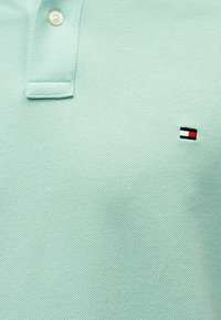 Ανοιχτό πράσινο πόλο από υφασμάτινη ύφανση με δύο κουμπιά στη λαιμόκοψη και μικρό κεντημένο λογότυπο σε κόκκινο, άσπρο και μαύρο.