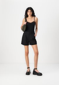 Svart tanktopp med blondekant, høy midje svarte shorts, og klumpete svarte sko. Bærer en grønn stoffveske. Glatte teksturer og moderne design.