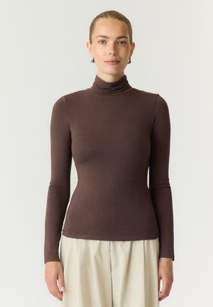 Femme aux cheveux tirés en arrière, portant un col roulé marron ajusté à manches longues et un pantalon beige ample, debout devant un fond uni.