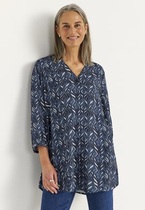 Lachende vrouw van middelbare leeftijd met grijs haar draagt een blauwe blouse met patroon en knopen en een blauwe spijkerbroek, staand tegen een witte muur.