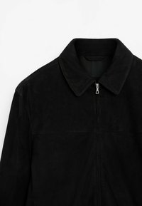 Veste en daim noire avec col et fermeture éclair argentée à l'avant, montrée de la poitrine vers le haut sur un fond blanc.