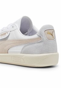 Sneakers Puma Palermo mają białą skórzaną cholewkę z beżowymi i szarymi zamszowymi akcentami, teksturowaną kremową gumową podeszwę oraz złote detale z logo.