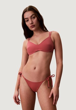 Młoda kobieta w różowym bikini z wiązanymi paskami na biodrach, stojąca na jednolitym jasnoszarym tle.