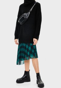Maglione oversize lavorato a maglia nero, gonna plissettata teal con motivo geometrico e stivali neri alla caviglia con borchie. Include una borsa a tracolla nera.