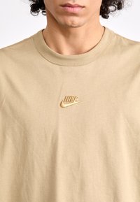 Bežinė medvilninė marškinėliai su apvalia iškirpte. Savybė - mažas siuvinėtas Nike logotipas šviesesnėje spalvoje ant krūtinės. Lygi tekstūra.