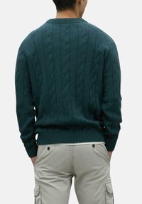 Suéter de punto color turquesa con patrón de trenzado y puños de canalé, visto desde la parte posterior, combinado con pantalones cargo de color claro.