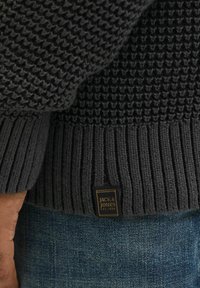 Dark gray knit sweater featuring textured patterns on the body and ribbed cuffs. Small black label with gold text at the lower hem.