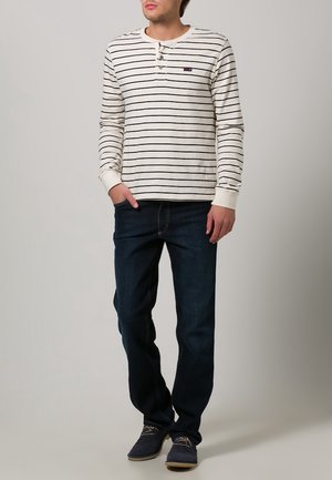 Camisa de rayas de algodón de manga larga en crema y azul marino, con cierre de cuello abotonado. Combinada con jeans azul oscuro y zapatos oscuros.