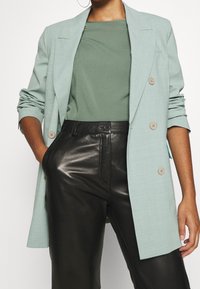 Světle modrý přizpůsobený blejzr s dvěma knoflíky, nošený přes zelený top, doplněný černými kalhotami z umělé kůže, zobrazuje elegantní siluetu.