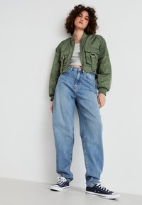 Chaqueta bomber verde oliva con dos bolsillos frontales, top gris corto, jeans sueltos de color azul claro, y zapatillas de color azul marino con cordones blancos.