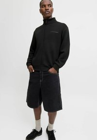 Ung mand iført en sort zip-up jakke, sorte knælange shorts, beige sokker og sorte sko, stående med hænderne i lommerne.