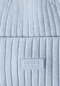 Ljust blå stickad mössa med ribbad textur, med en rektangulär gummipatch märkta "FÄRGFULLA SIDOR AV LIVET" på framsidan.