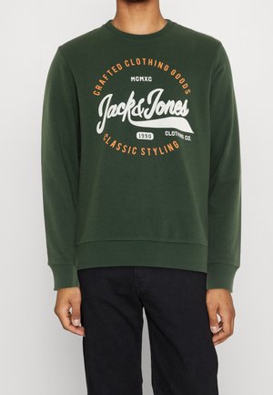 Osoba ubrana w ciemnozieloną bluzę z pomarańczowo-białym logo Jack & Jones i czarne spodnie na jednolitym tle.