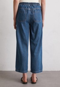 Blauwe denim wijde broek met een elastische tailleband, voorzien van twee achterzakken en ruwe zomen, tegen een neutrale achtergrond geplaatst.