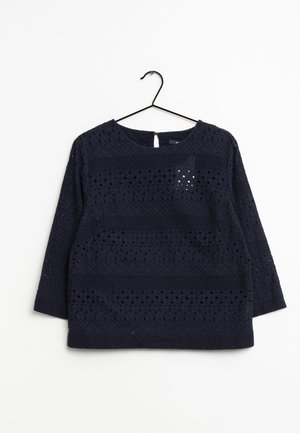 Marineblauwe blouse met lange mouwen en ajourkantpatroon, ronde halslijn, keyhole knoopsluiting, hangend aan een zwarte metalen hanger op een witte achtergrond.