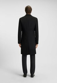 Cappotto di lana nero con un design su misura, caratterizzato da una silhouette lunga, colletto a revers e polsini con bottoni, indossato sopra pantaloni coordinati.