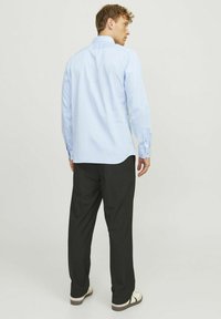 Chemise bleu clair à manches longues avec un ourlet arrondi et des poignets boutonnés, associée à un pantalon noir et des sandales blanches, vue de dos.