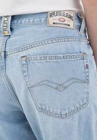 Nära bild av ljusblå Replay Blue Jeans bakficka med fint broderi och varumärkesetikett på midjebandet.