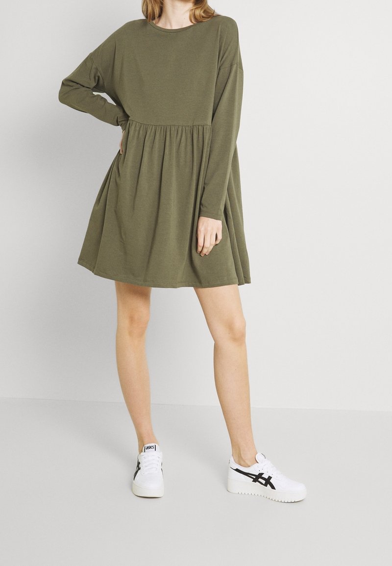 Robe longue olive à manches longues avec une taille empire froncée. Associée à des baskets blanches avec des accents noirs et une semelle texturée.