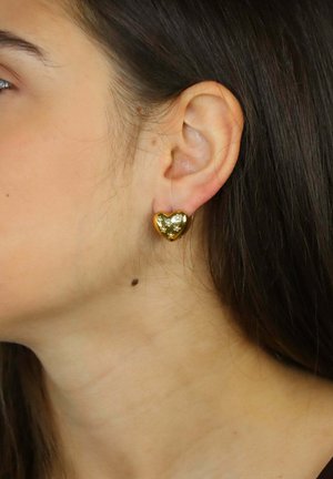 Złote kolczyki w kształcie serca z błyszczącym wykończeniem i subtelnymi gwiazdkowymi akcentami, noszone na uchu. Zbliżenie podkreślające ich design.