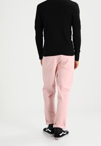 Persoon in een zwart langmouwshirt, lichtroze broek en zwart-witte sneakers, staande met de rug naar de camera op een witte achtergrond.